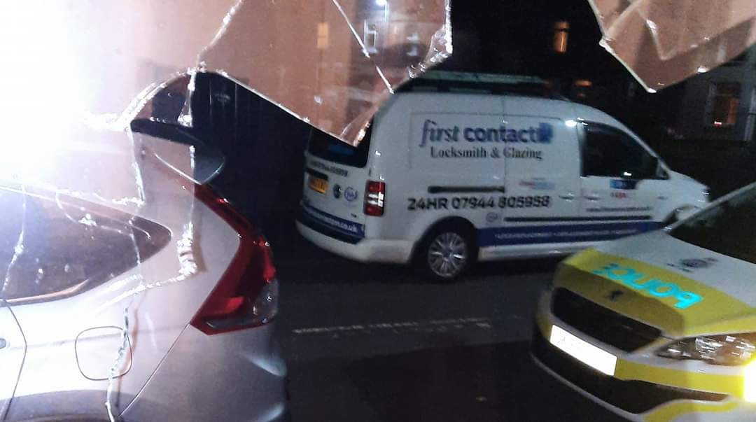 A night call-out seen through a smashed pane of glass: the First Contact van parked in the street beside a police car
