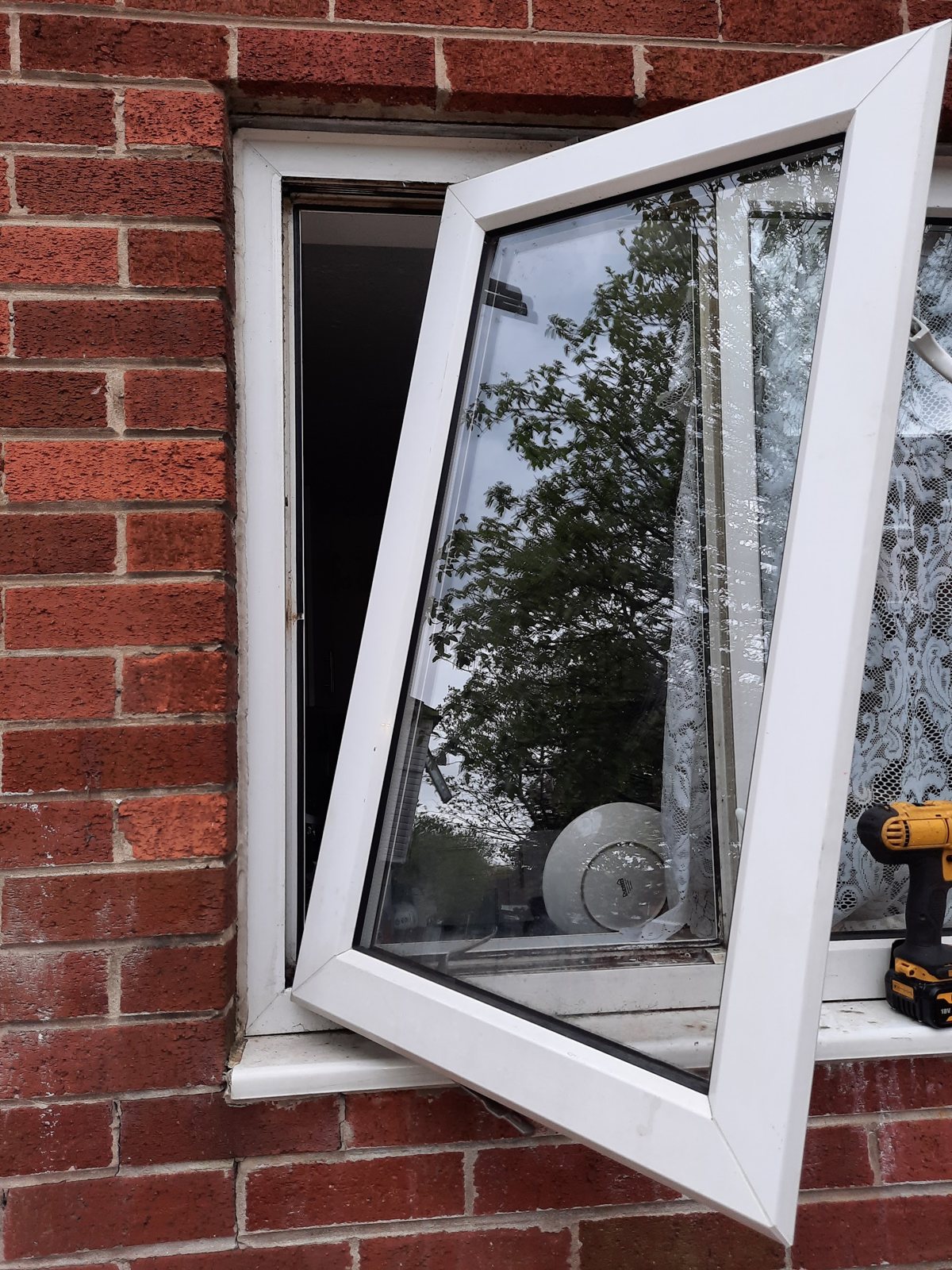 A white uPVC window swung open from its frame in a brick wall during a repair, a cordless drill on the sill